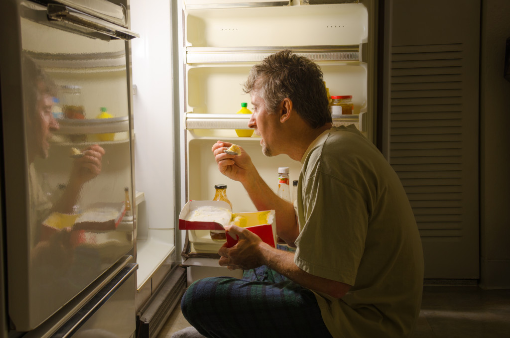 A man who has nighttime sleep-related eating disorder sleep eating as he sits in front of a refrigerator eating ice cream out of carton