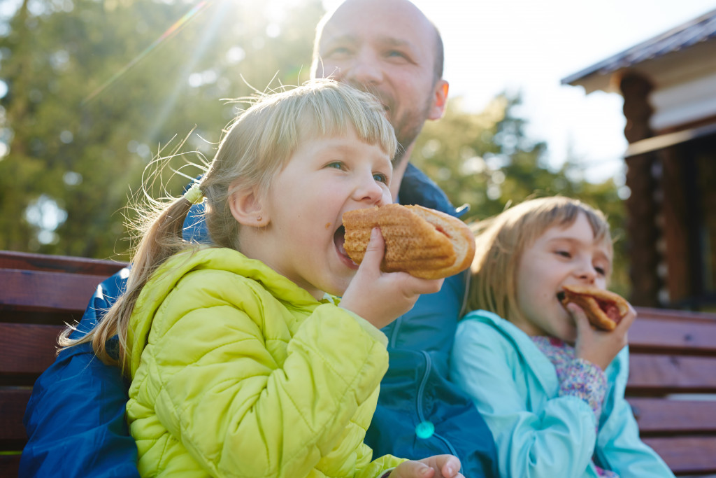 A picture of two children eating hotdogs on buns, together with their father.